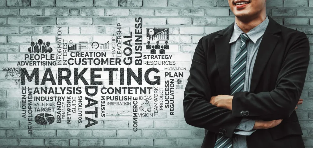 Businessman standing with arms crossed in front of marketing-related word cloud on a brick wall.